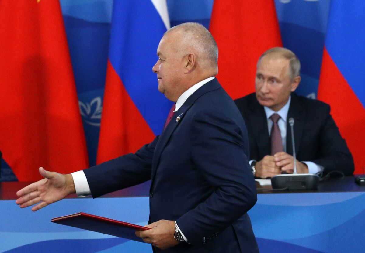 A man in a suit reaches out his hand. Behind him is another man seated in front of red, blue and white flags