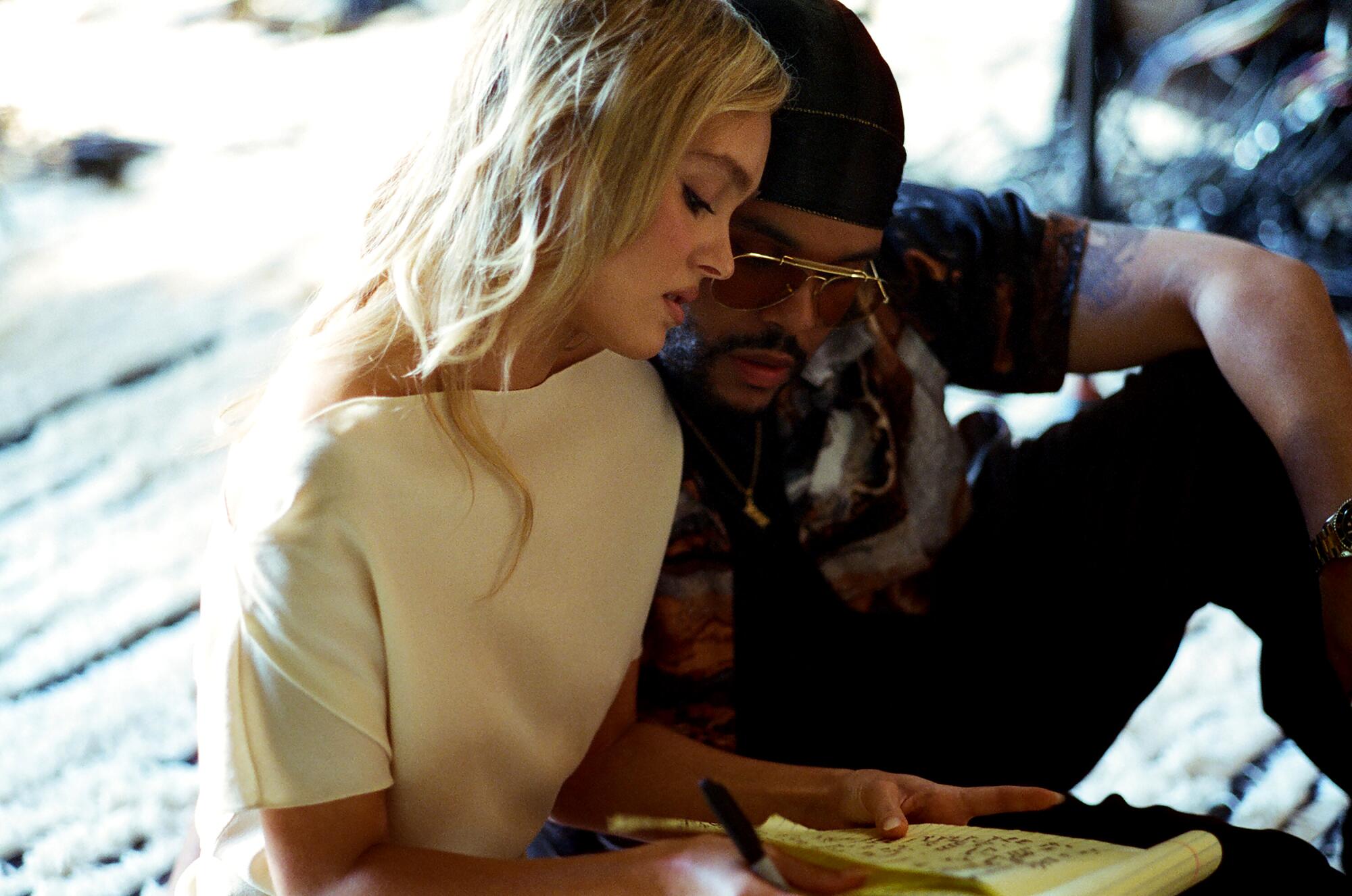 A blonde woman in a white shirt looks at a notebook and a man in dark clothing leans against her left shoulder.