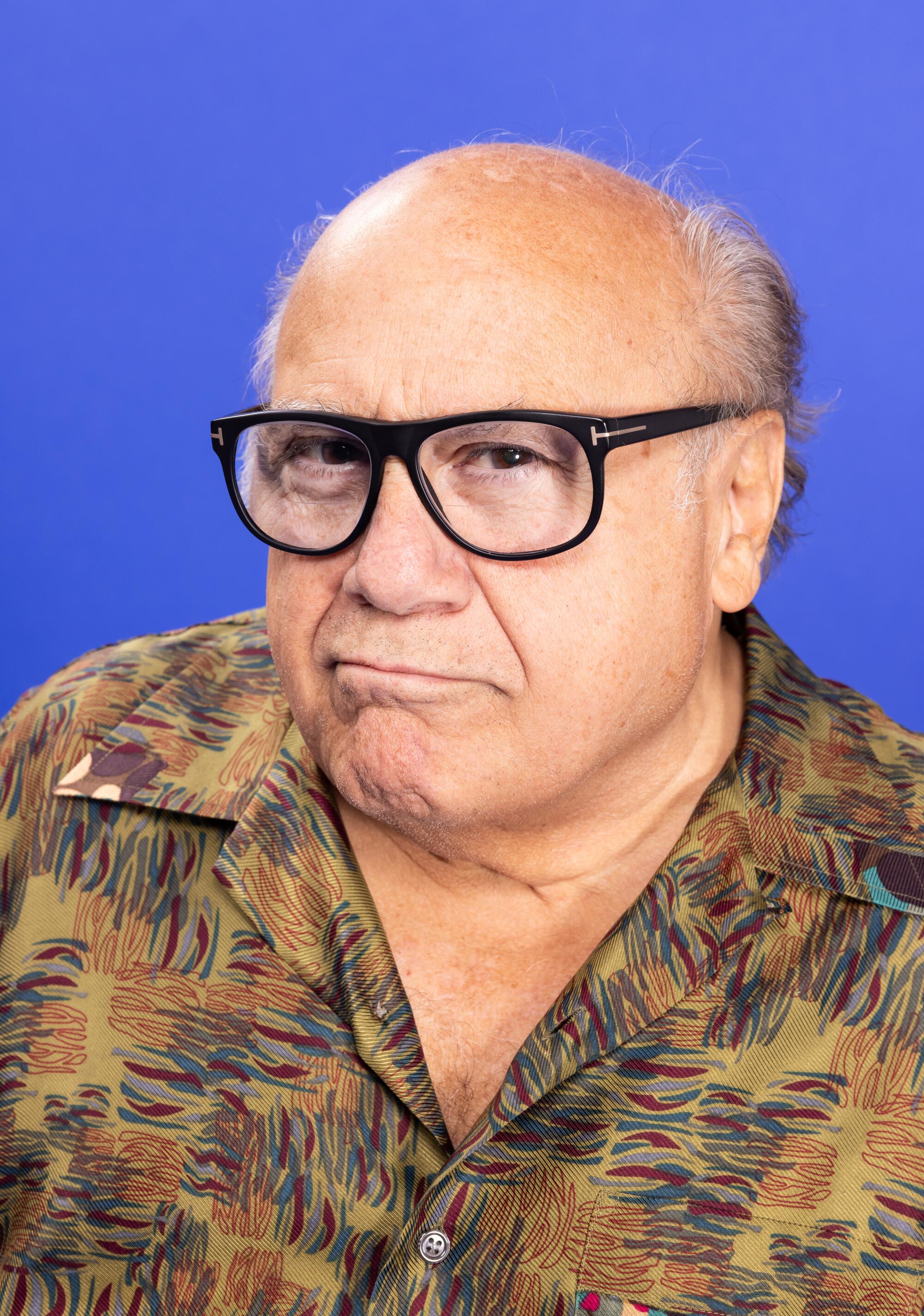 A bald man with a slight scowl in black-rimmed glasses in a brown patterned shirt.