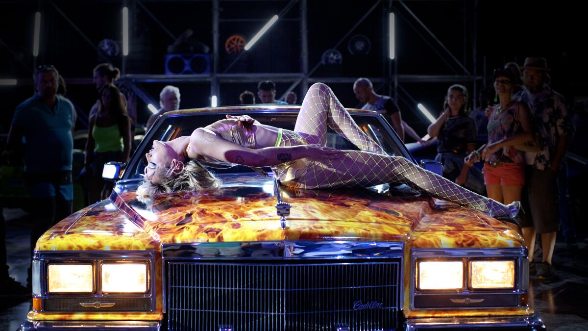 A woman in fishnet stockings and a bikini top lies on top of a car with painted flames