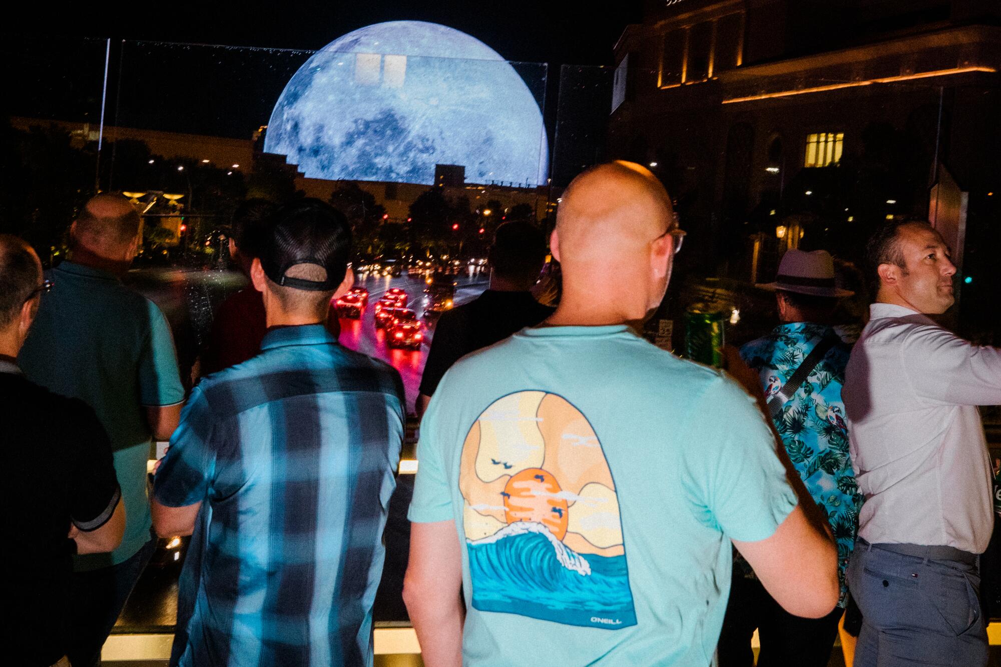 People view a spherical building broadcasting images of the moon from a pedestrian bridge over a busy street.