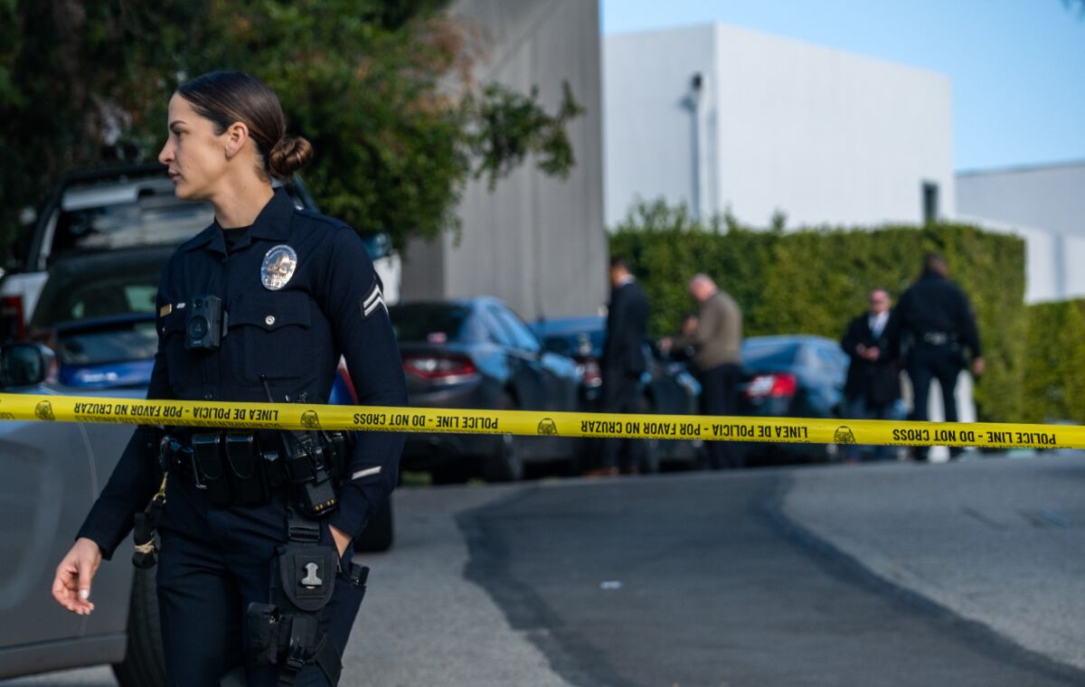 An L.A. police officer stands near yellow crime tape with investigators in the background.