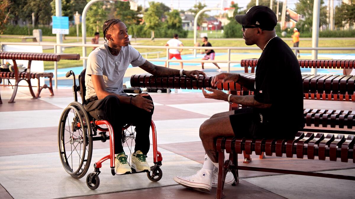 A man in a wheelchair listens to a man talking to him from a bench.