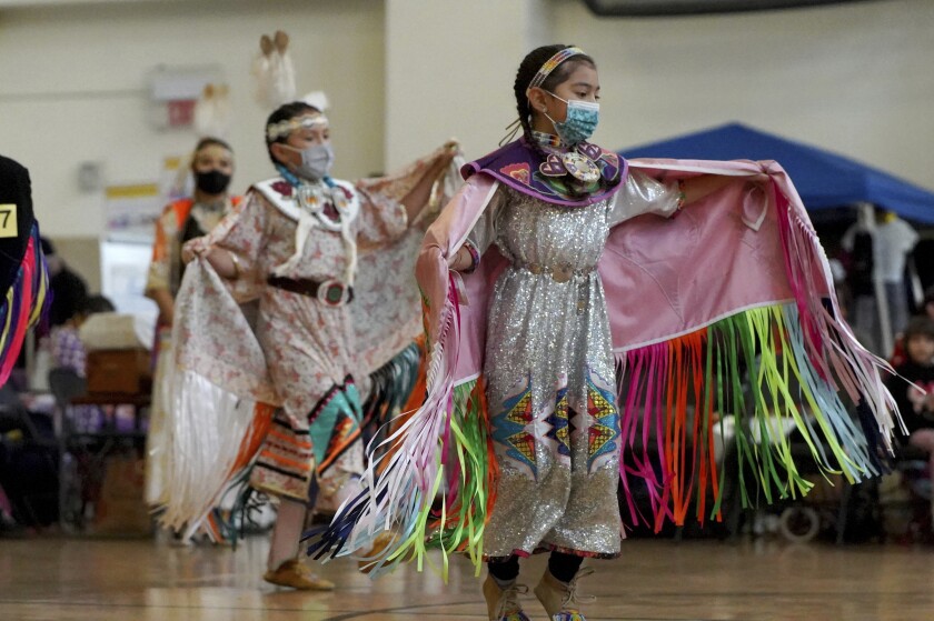In this photo provided by the American Indian Child Resource Center, Native American children dance while wearing masks at a powwow hosted by the American Indian Child Resource Center featuring traditional Native American music and dancing, along with the chance to get COVID-19 vaccinations and testing, on Dec. 4, 2021, at La Escuelita Elementary School, in Oakland, Calif. (Garrett Rich/American Indian Child Resource Center via AP)
