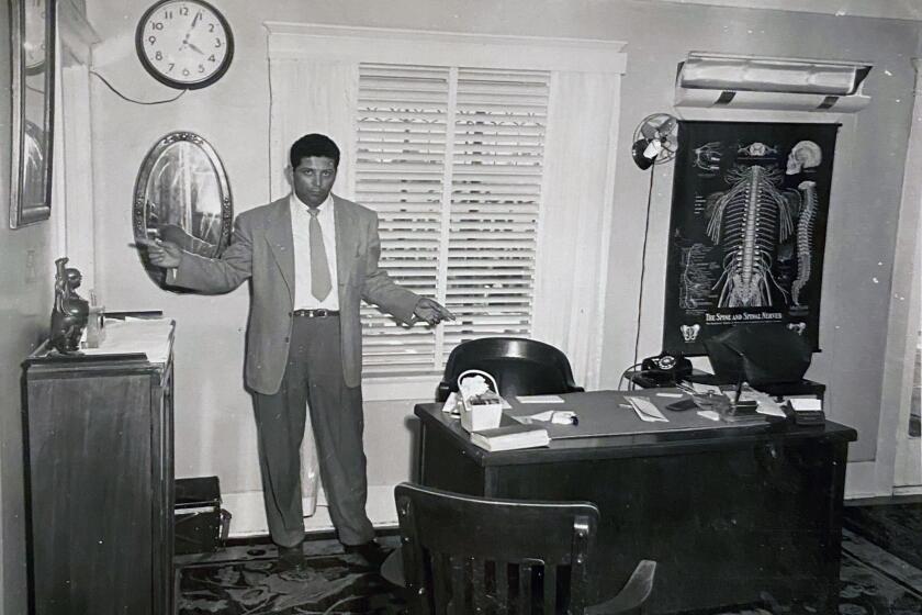 Danny Galindo wearing a 1960s-style light suit and light tie standing beside a desk and other bedroom furniture.
