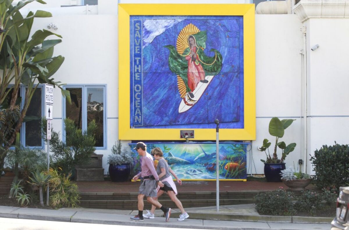 Pedestrians walk past the “Surfing Madonna” mosaic displayed on Encinitas Boulevard, near the Coast Highway intersection, on Dec. 24, 2019 in Encinitas.