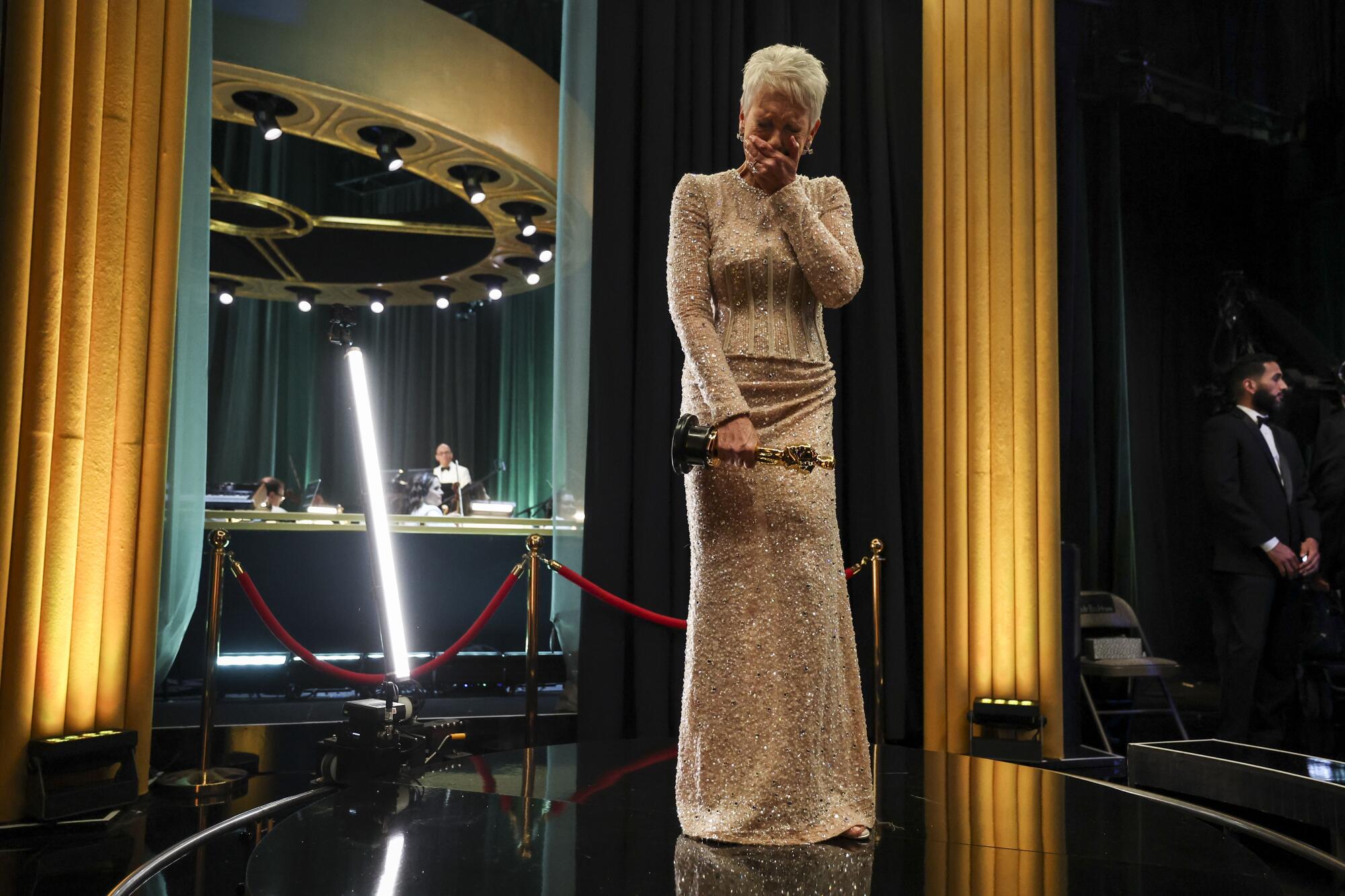 Backstage, a woman in a long gown has one hand on her Oscar statuette and the other hand over her mouth as she cries in joy.