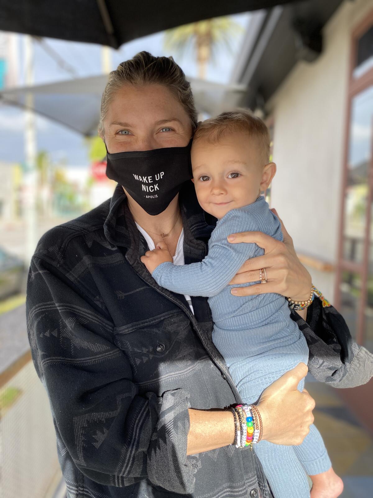 Amanda Kloots, wearing a "Wake Up Nick" mask, holds baby Elvis.