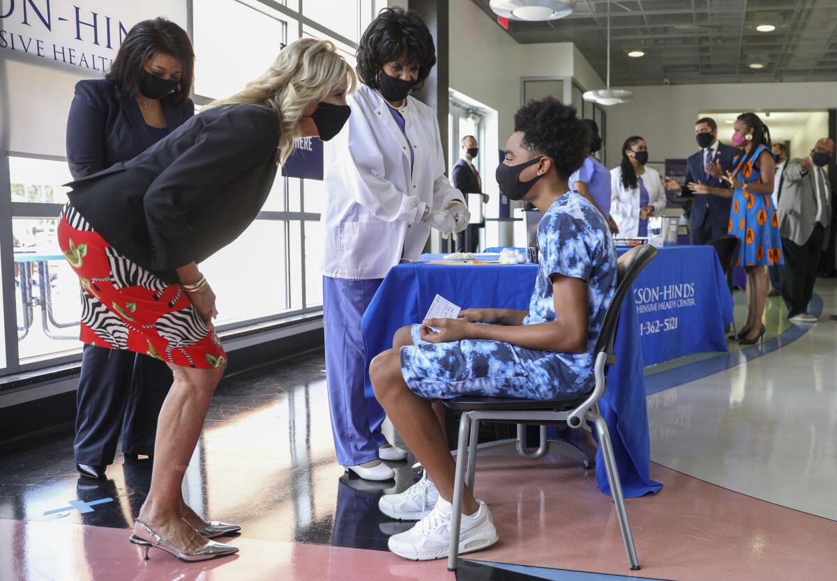 First Lady Jill Biden, in a black mask, leans down to speak to a masked young man seated near health workers