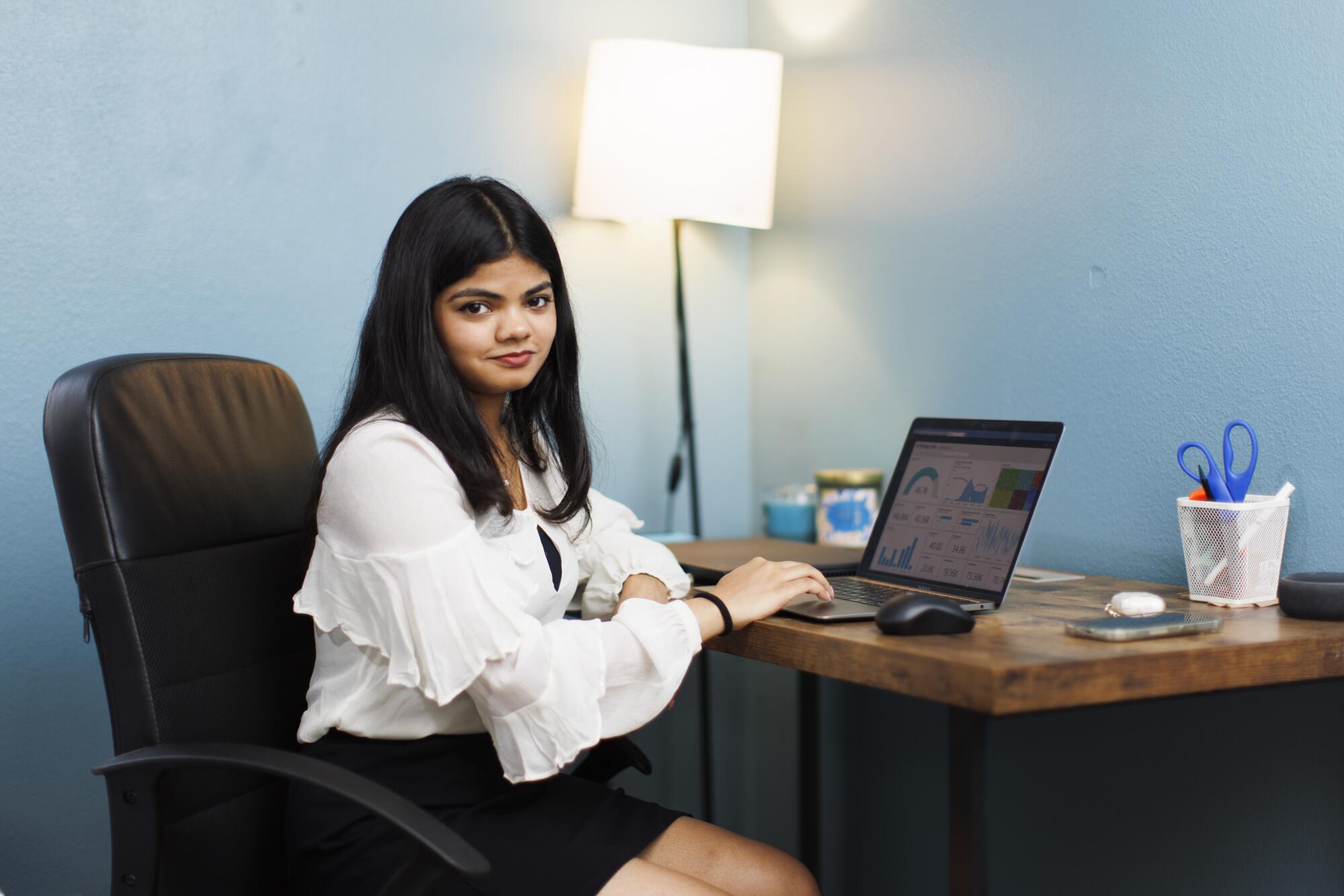 A woman in a white blouse sits at a desk with a laptop.