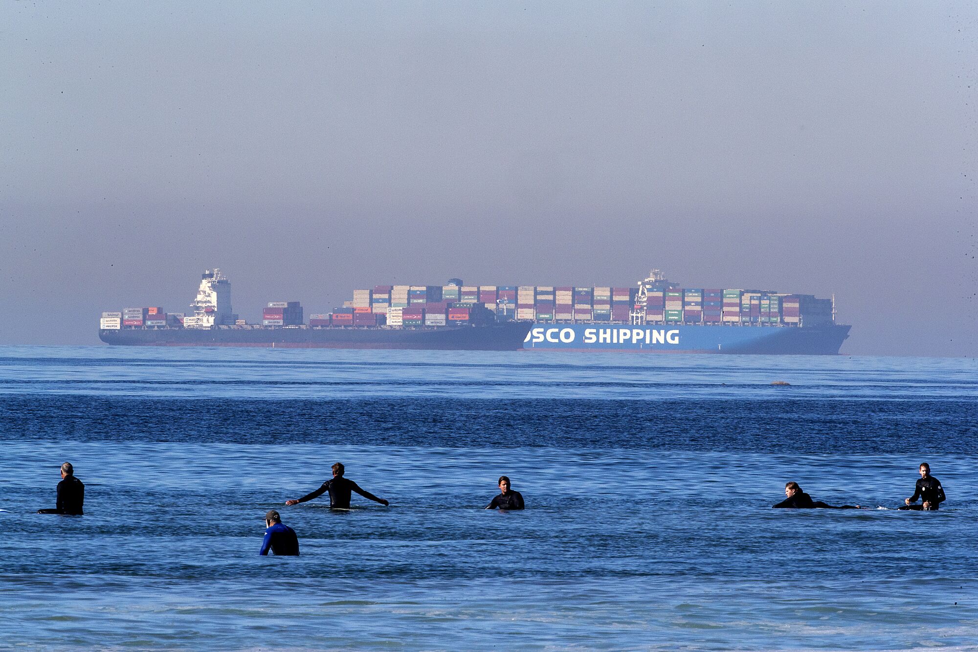 Surfers in black wetsuits bob in the water. In the background is a container ship. 