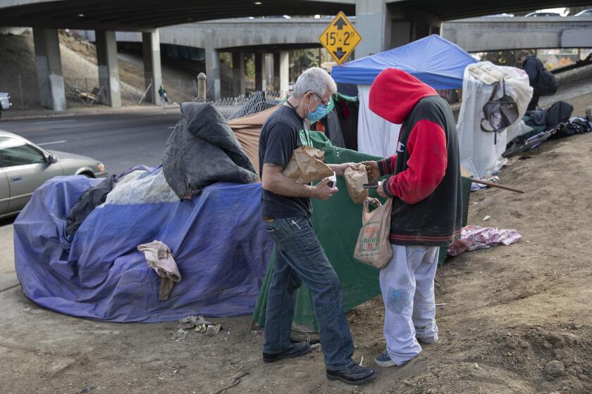 Hollywood, CA - December 10, 2021:Jason Sodenkamp, left, a case manager for Community Health Project LA, hands a care package containing food, hygiene products and water to a client who goes by the nickname, "Lucifer," at a homeless encampment on Virgil Ave. near the 101 freeway in Los Angeles. (Mel Melcon / Los Angeles Times)