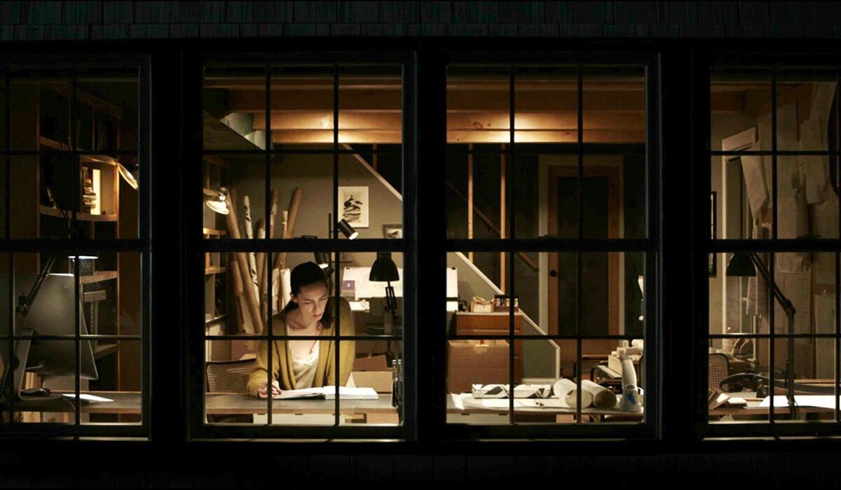A woman seen through a window reading a book in the movie "The Night House."