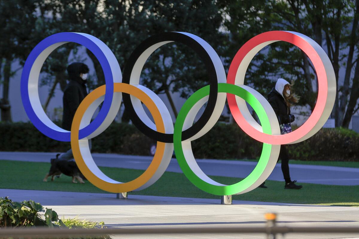 People wearing a face mask walk by the Olympic rings  