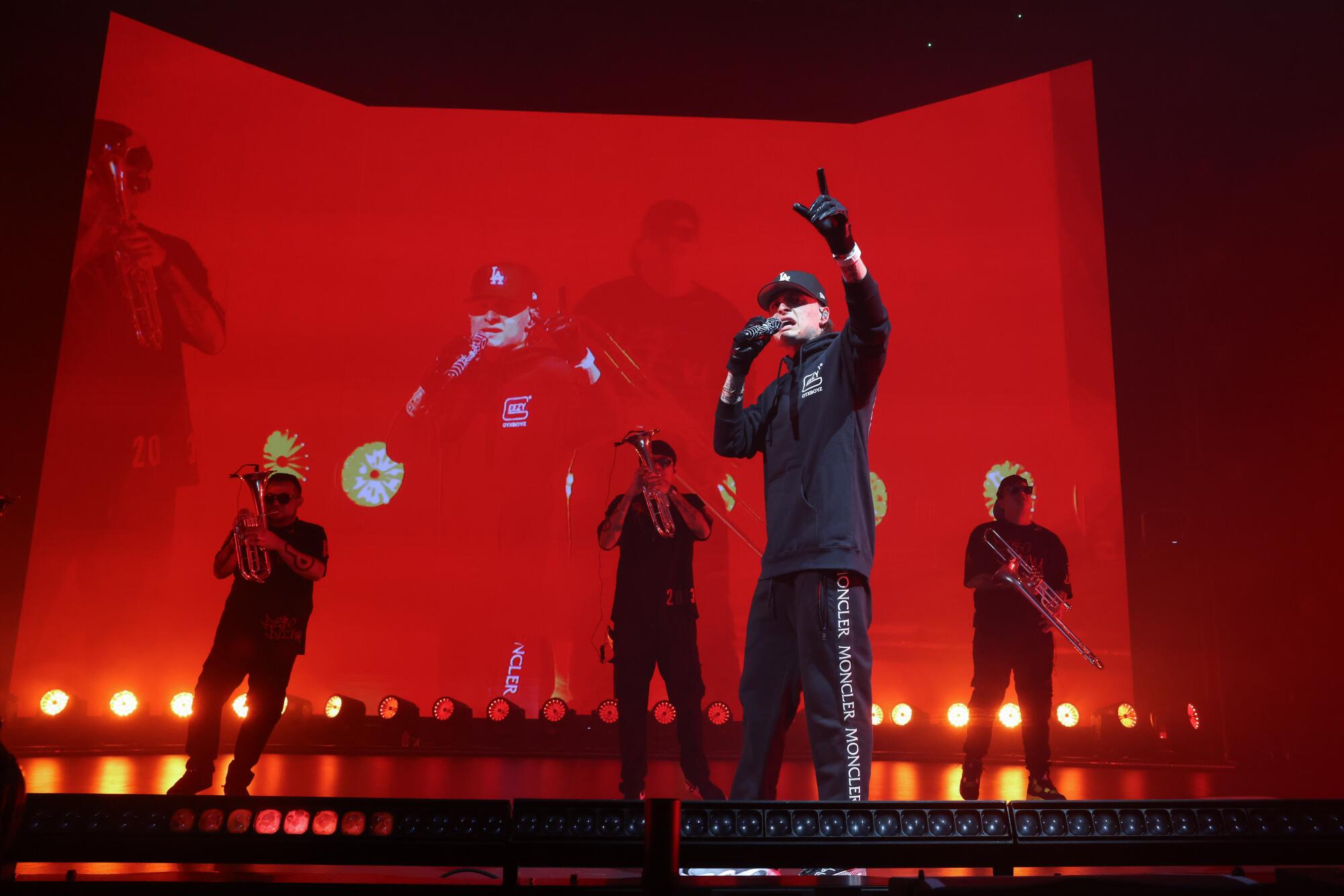 Peso Pluma in a black outfit with musicians playing behind him on a stage with a red screen backdrop.