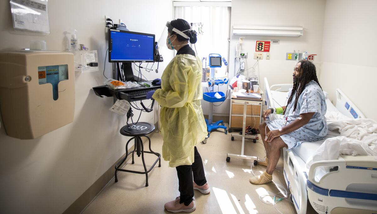 A woman in a mask and face shield stands before a computer screen. Behind her, a man in seated on a hospital bed