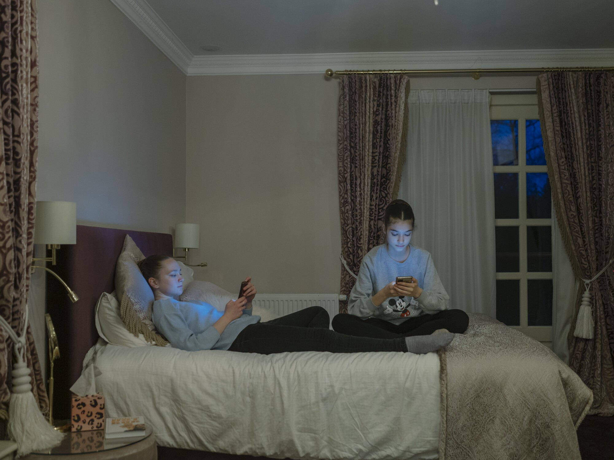 Two girls sitting on a bed looking at their phones 