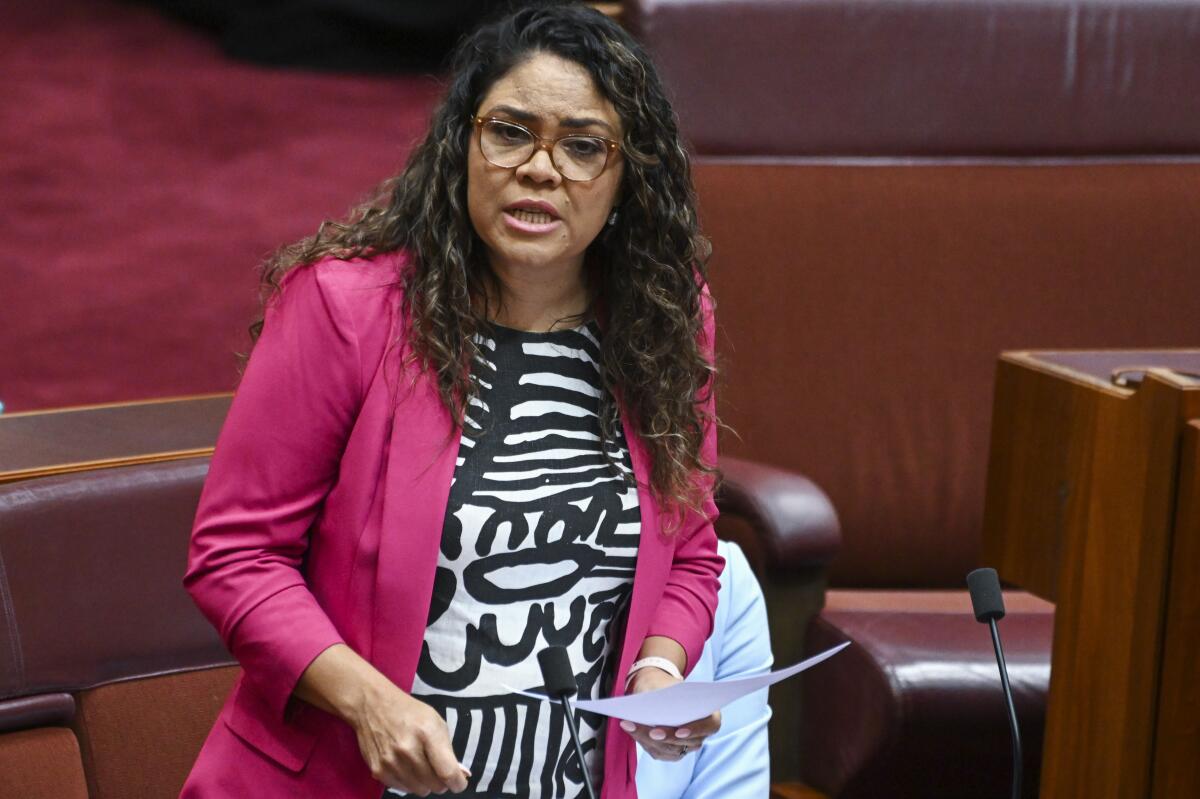 A woman with long dark hair, in glasses, pink jacket and a patterned top, holds a document while speaking 