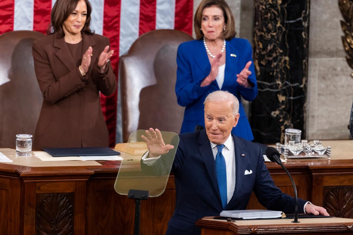 President Biden waves at a lectern with Vice President Kamala Harris and House Speaker Nancy Pelosi behind him. 