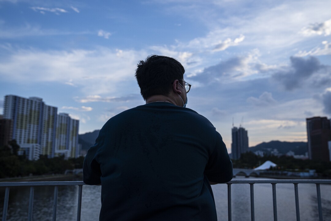 Seen from behind, a man leans on a bridge railing in Hong Kong looking out over the river and high-rise buildings.