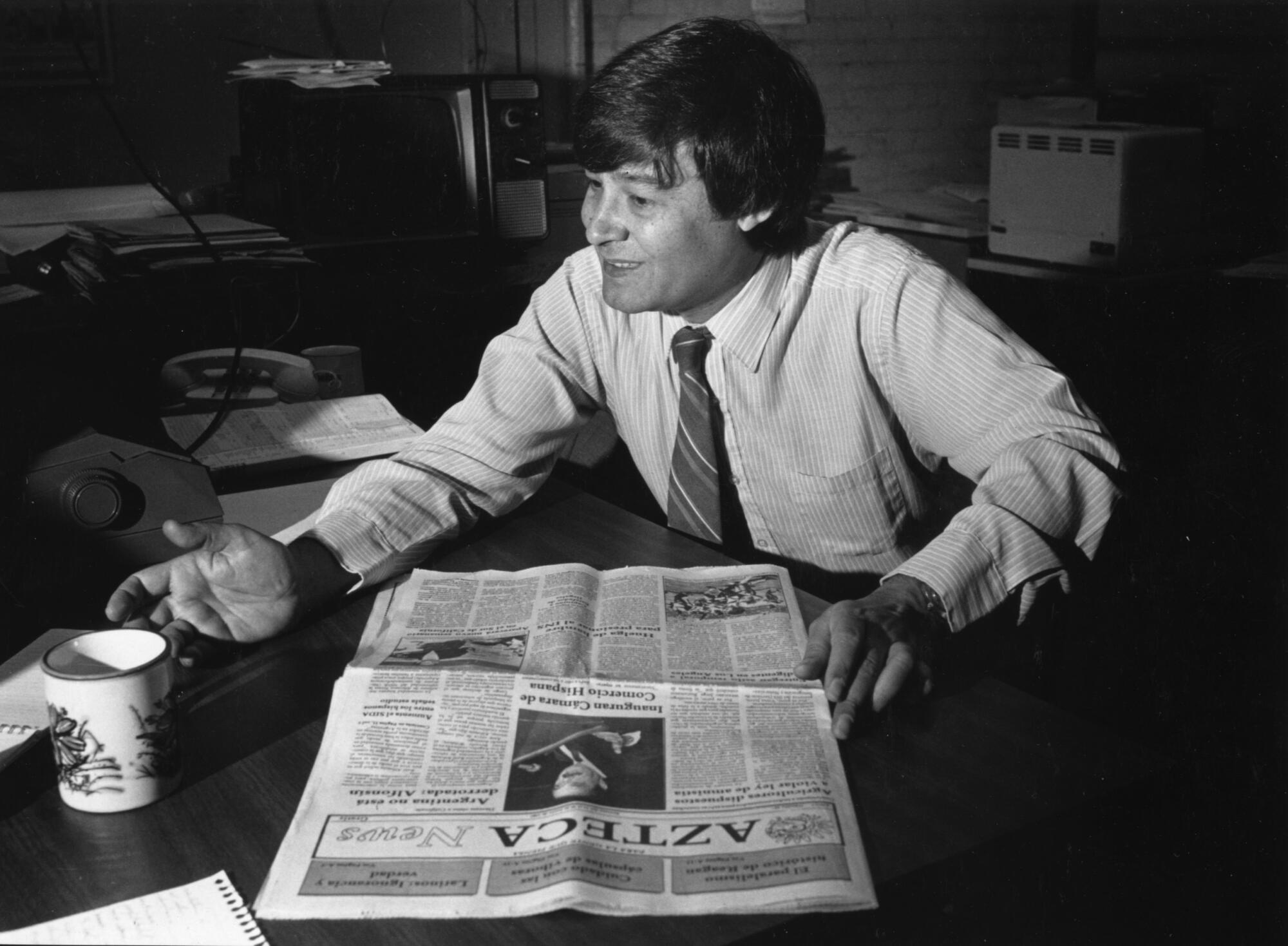 A black and white photo shows a seated man with a newspaper in front of him on a table.