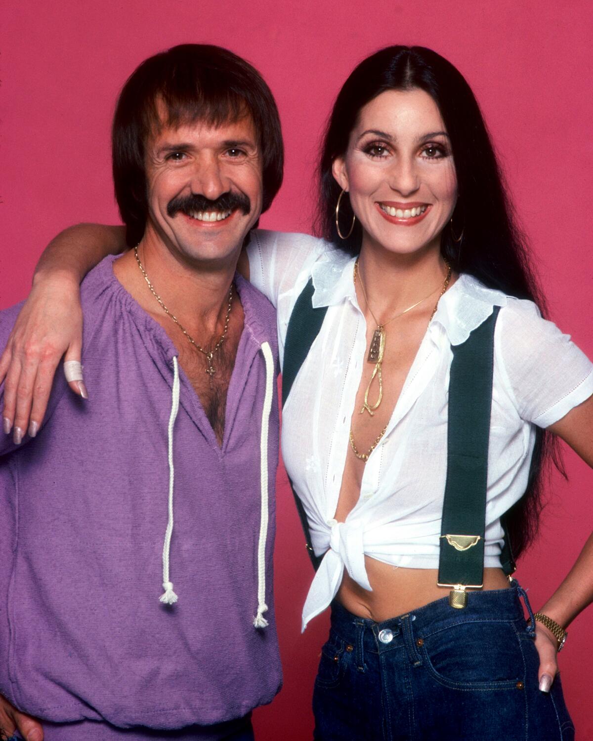A man with long hair and a mustache in a purple top and a woman with long hair in a white top with suspenders