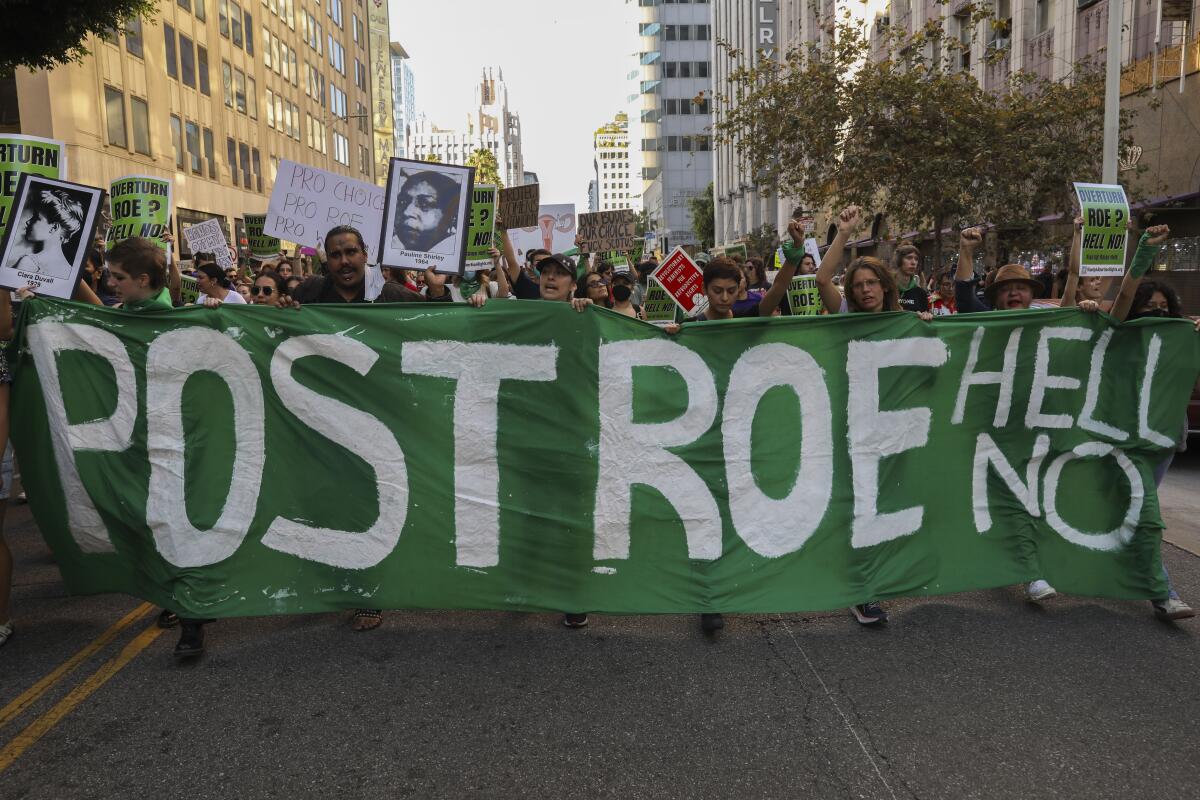 A crowd of protesters march in a street carrying a green banner that says "Post Roe, Hell No"