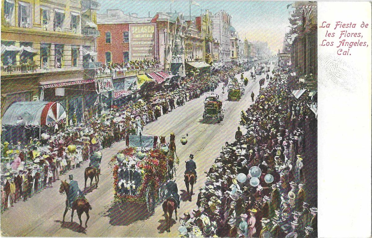 Horse-drawn carriages decked with flowers parade along a downtown street, with crowds on either side.