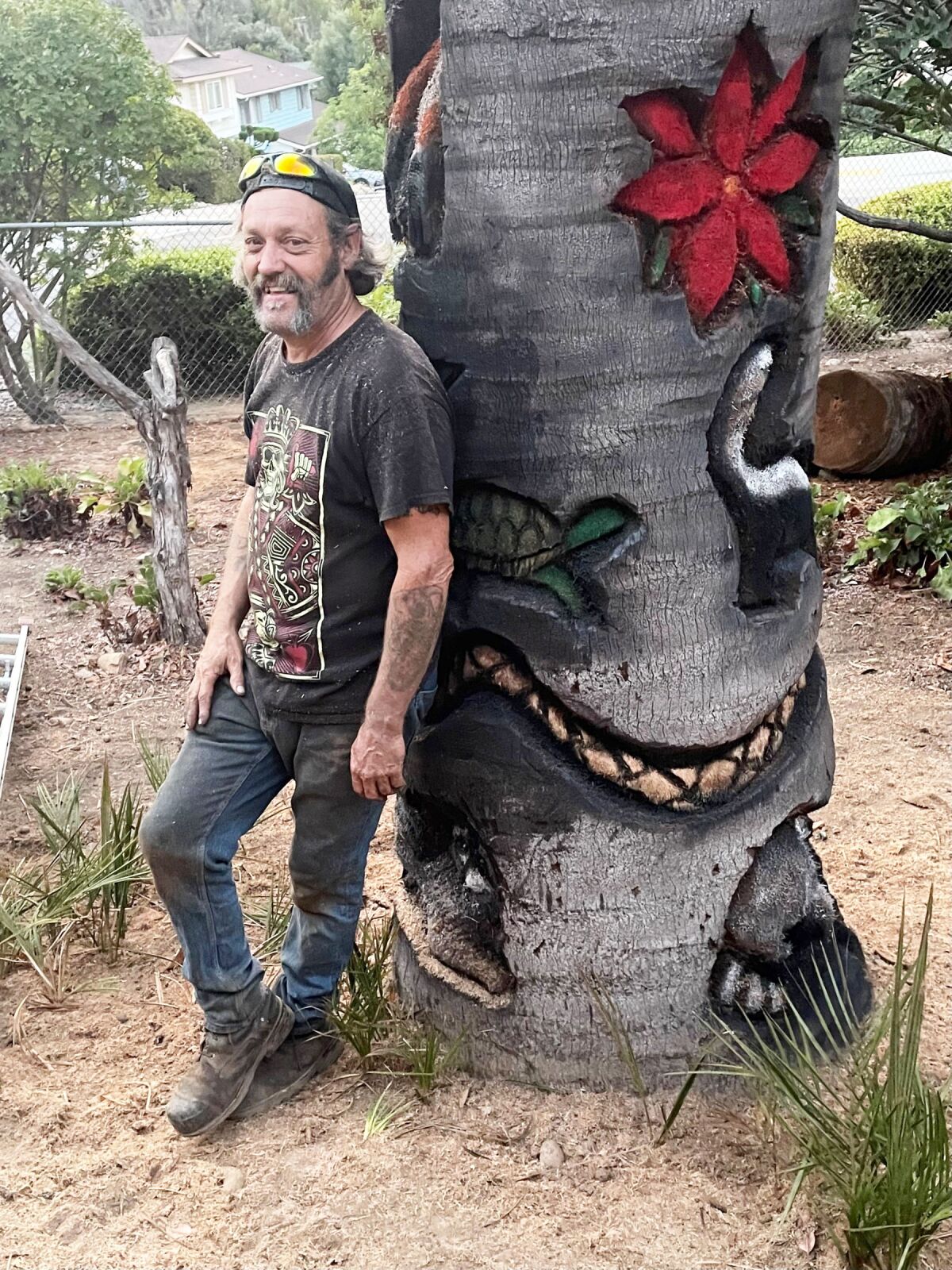 Dan Bialk, aka “Tiki Dan,” poses beside his artwork on the trunk of Carol Mulcahy’s palm.