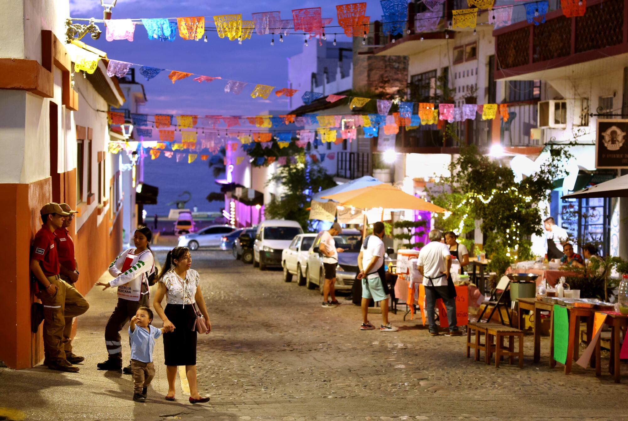People linger, walk and dine along a dirt street decorated with banners in several colors strung overhead