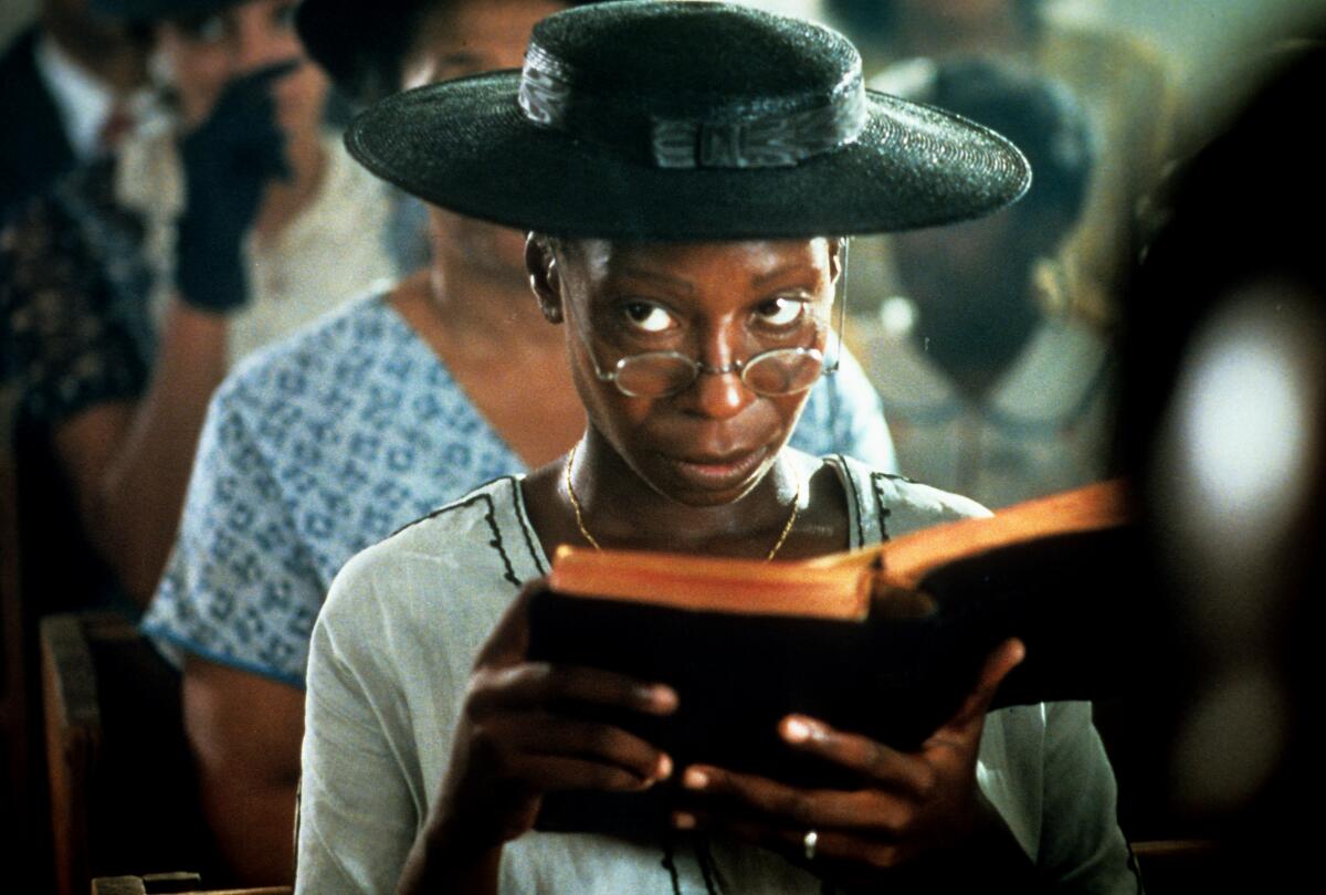A woman sits in church, holding an open book, wearing a hat and peering over her glasses