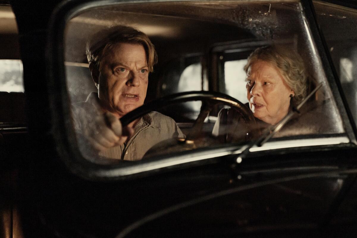 Eddie Izzard, alongside Judi Dench, drives a car.