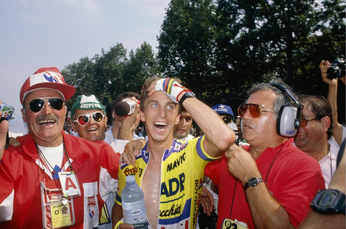 A cyclist in a yellow jersey looks gleeful. A broadcaster in red shirt with headphones and a mic stands next to him.