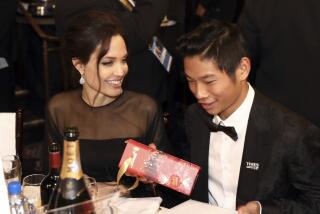 Angelina Jolie in a black mesh dress smiling and sitting next to son Pax who wears a tuxedo and holds a bar of chocolate