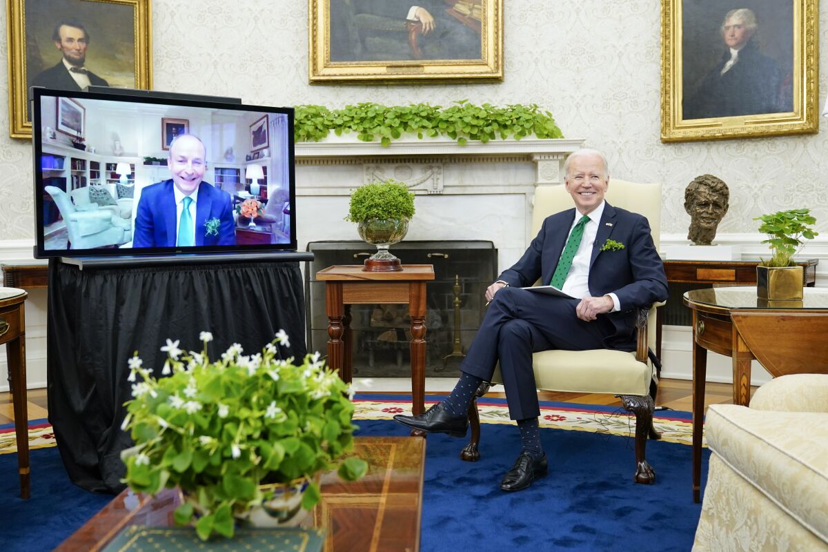 A man in a suit and green tie smiles as he sits before a screen showing another smiling man, also in suit and green tie 