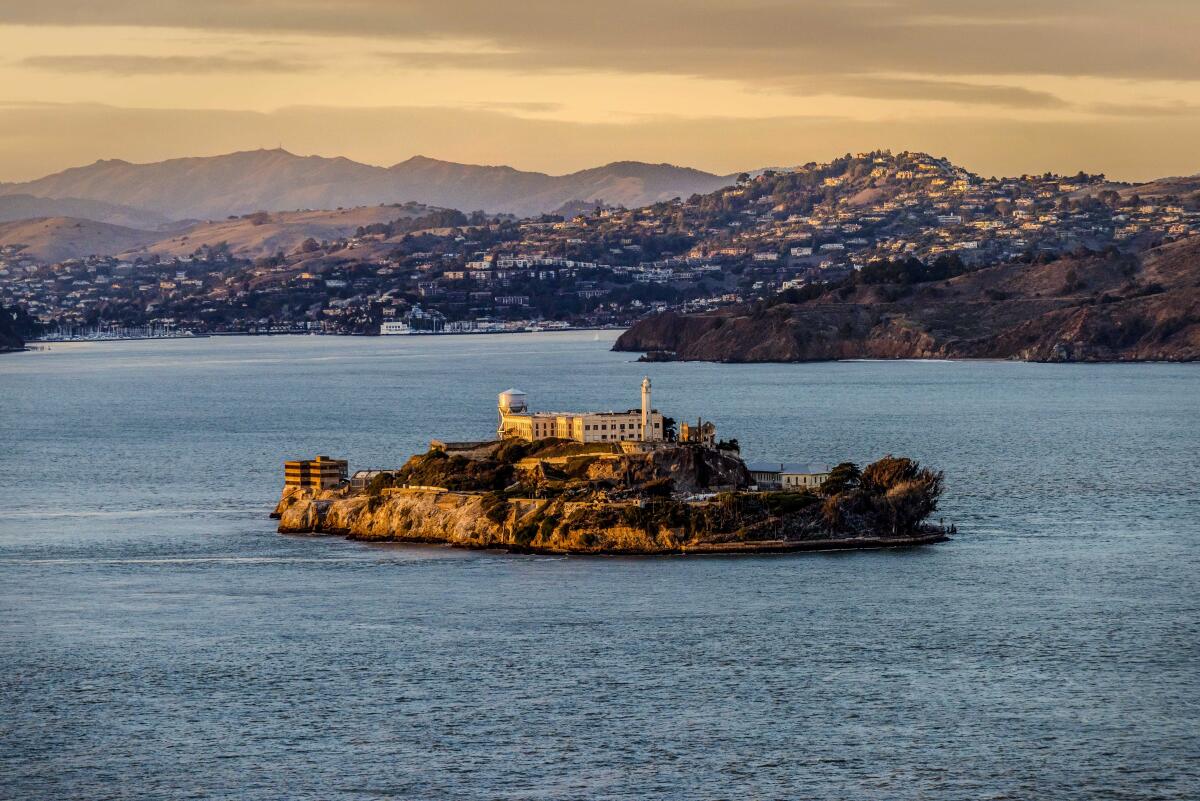 From a distance, a small island with a multi-story building is seen. In the background are hills with trees and homes.