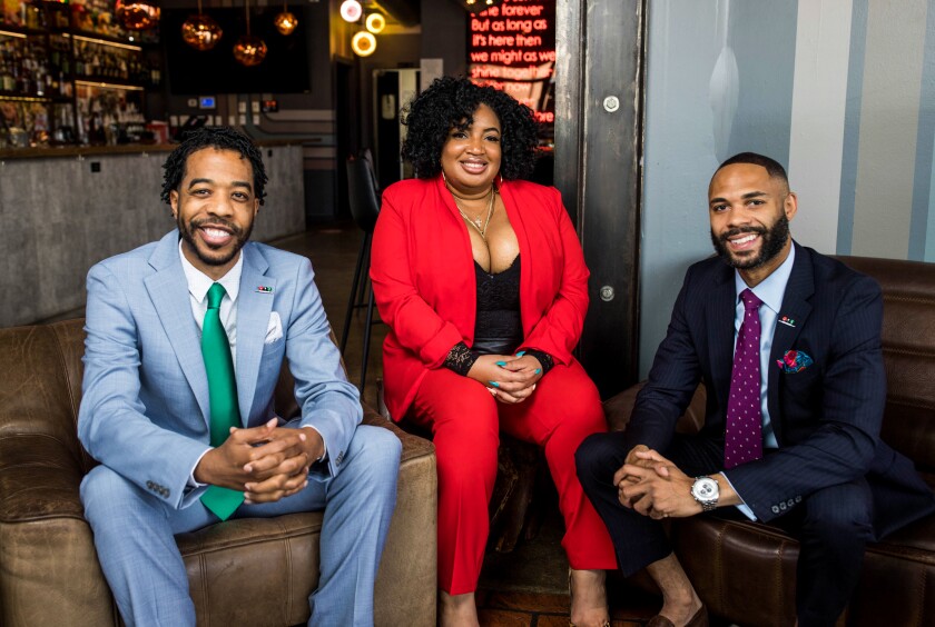 A woman in a red pantsuit sits between a man in a baby blue suit and green tie, left, and a man in a black suit and tie.