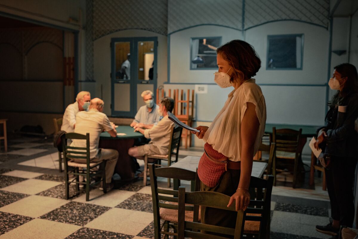 A woman reading a script wears a mask while four men in the background sit at a table.