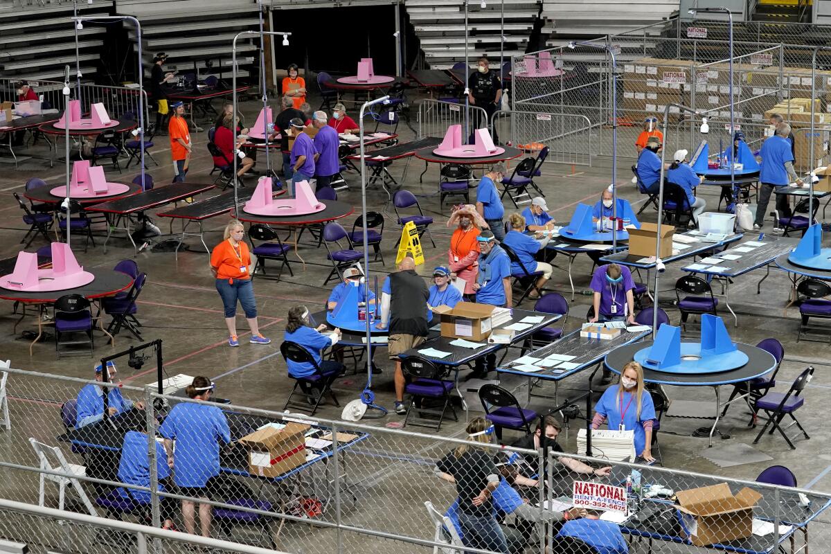Ballots are examined and recounted by several people in different colored shirts at tables behind chain link fence.