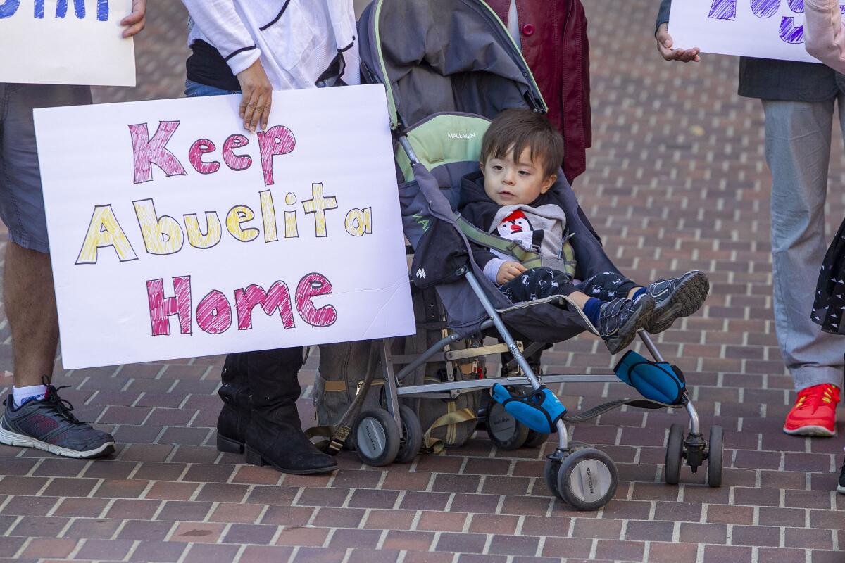 A toddler sits in a stroller next to a woman holding a sign that says "Keep abuelita home."