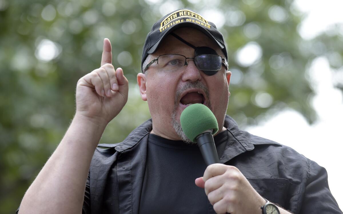 A man in a cap that reads "Oath Keepers lifetime member" pointing a finger up as he speaks into a microphone outdoors.