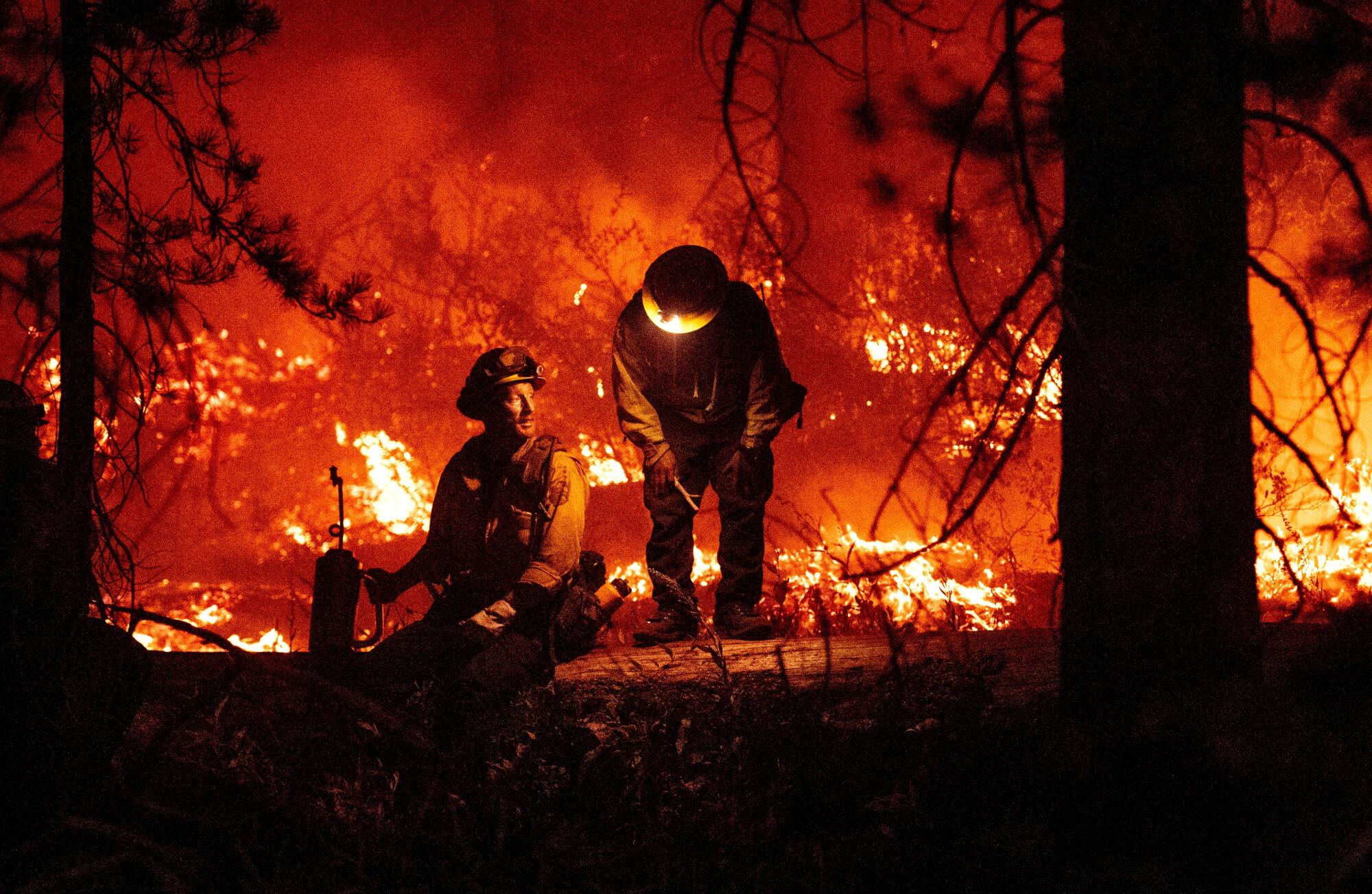 A firefighter sits on a log with another standing. Flames rage in the background. 
