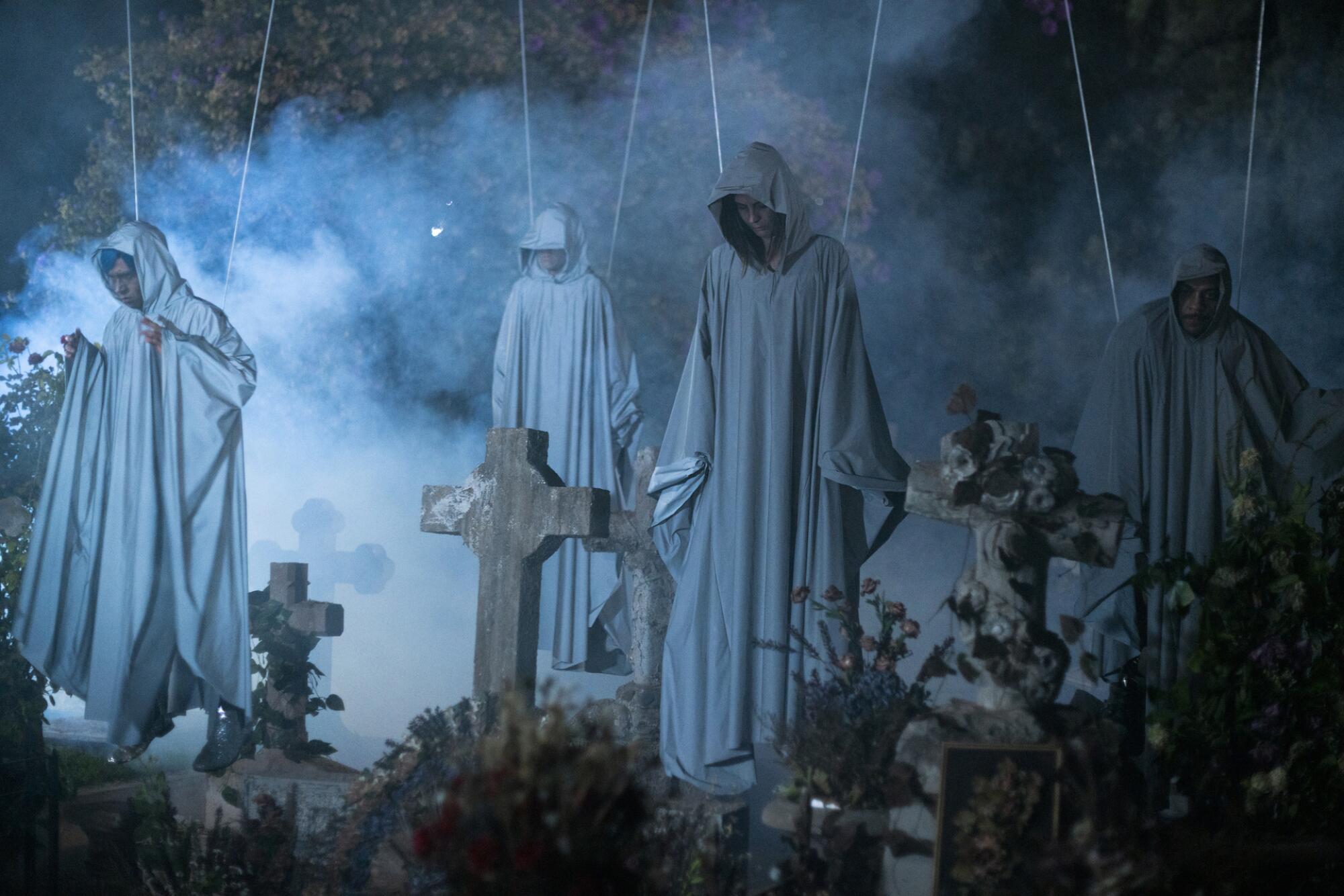 Four people in ghost outfits are seen hanging from wires above a cemetery.