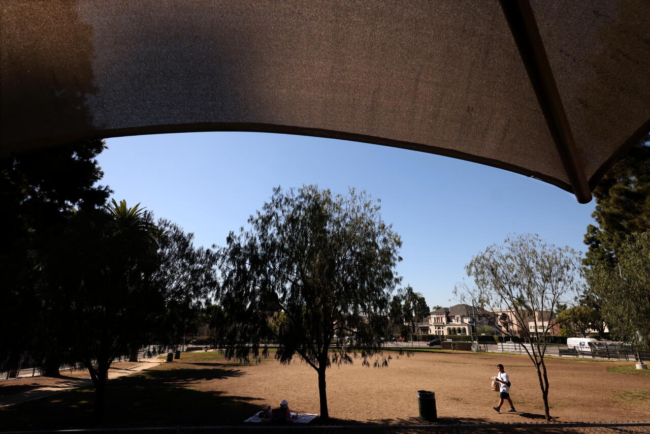 Some fabric shade hangs over an area in the foreground of a park. A man is walking in the distance.
