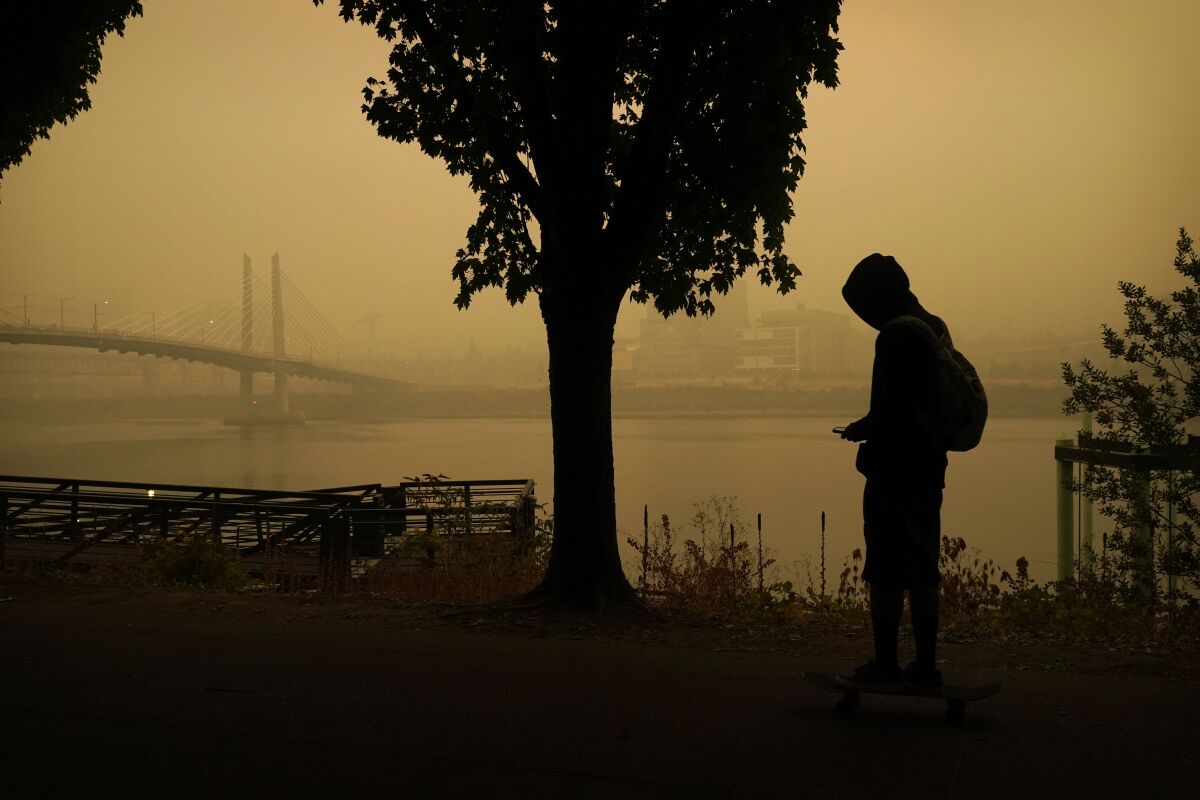 A person rides a skateboard along the Willamette River as smoke from wildfires partially obscures the Tilikum Crossing Bridge, Saturday, Sept. 12, 2020, in Portland, Ore. (AP Photo/John Locher)