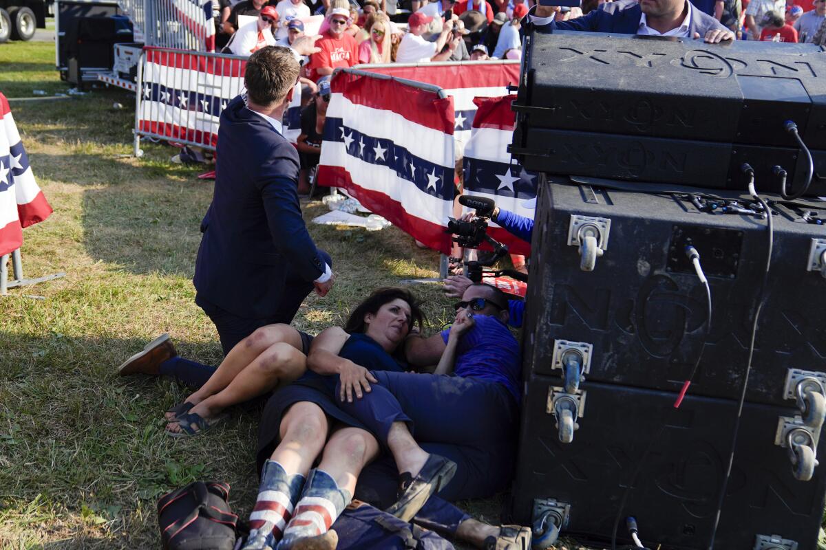 Civilians take cover on grass near sound equipment and American flags as a Secret Service agent in a suit kneels nearby.