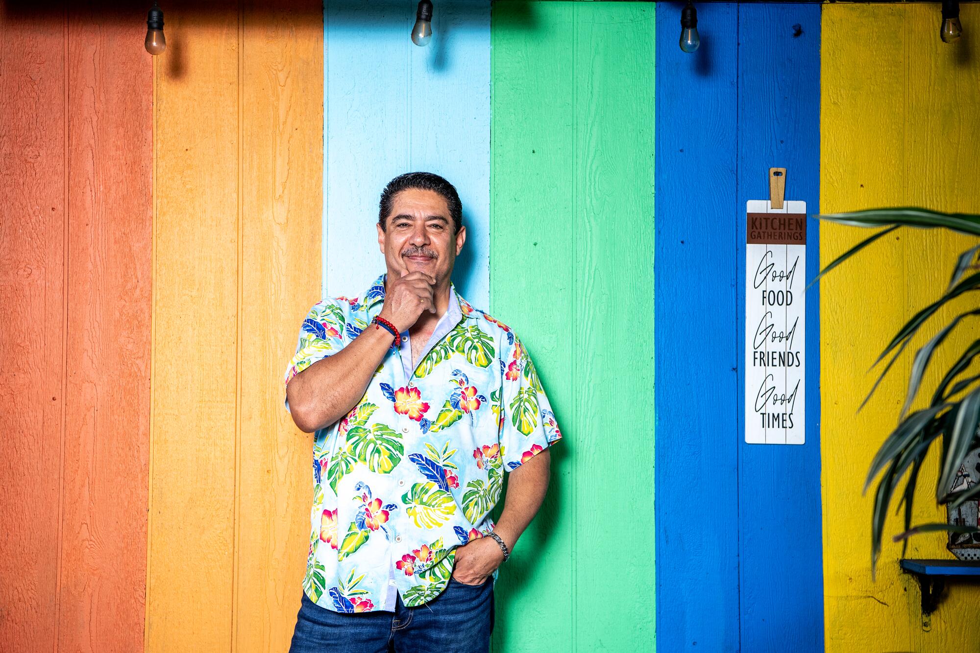 A man in an aloha shirt stands in front of a wall painted in colorful vertical stripes