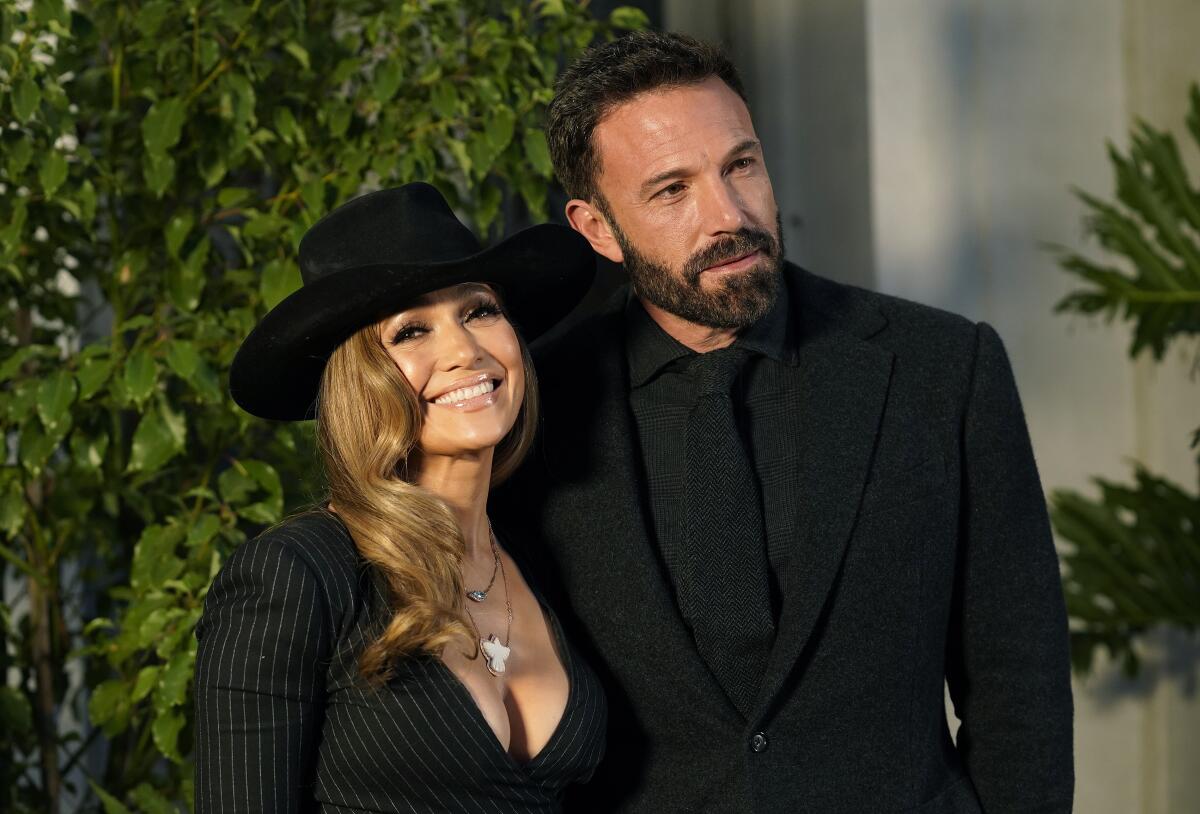 Jennifer Lopez in a black wide-brimmed hat and a black dress with a deep neckline next to Ben Affleck in a black suit