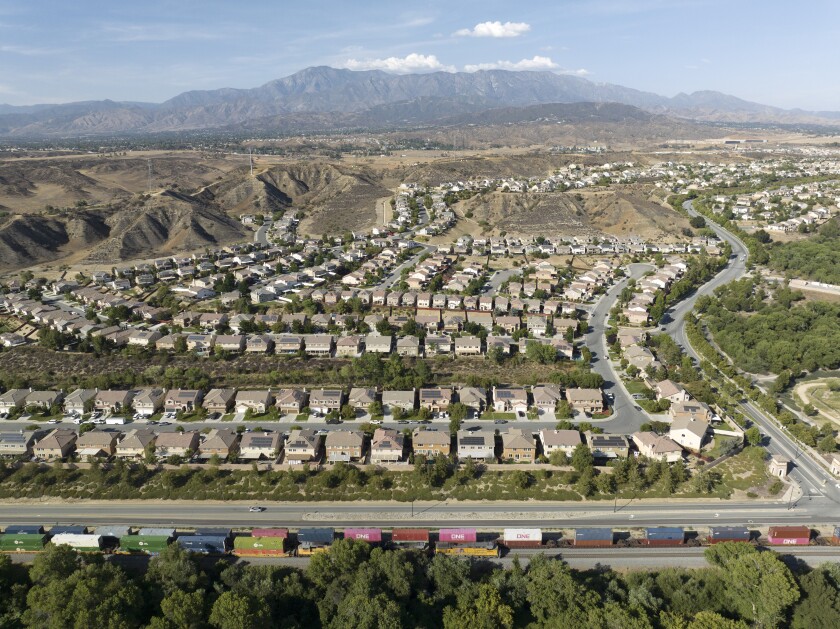 An aerial view of a housing development backed by mountains. A train passes along in the foreground.