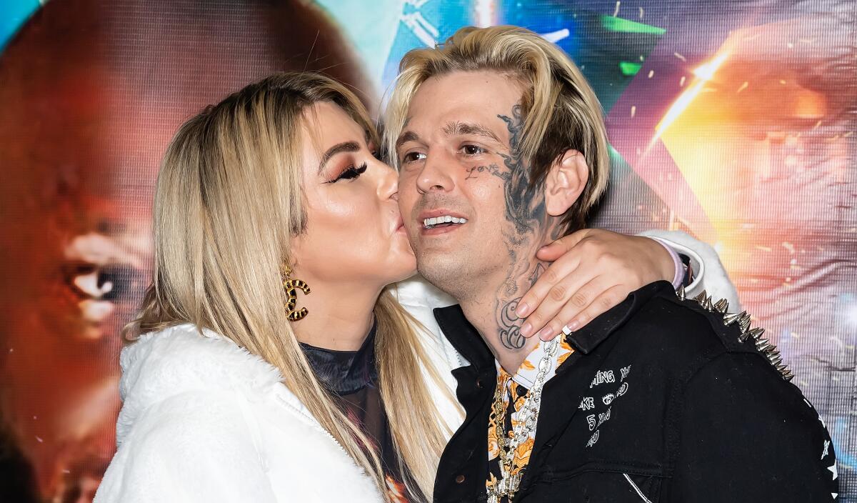 A blond woman in a white coat kisses a blond man with face tattoos on cheek and neck while holding her arm around his neck